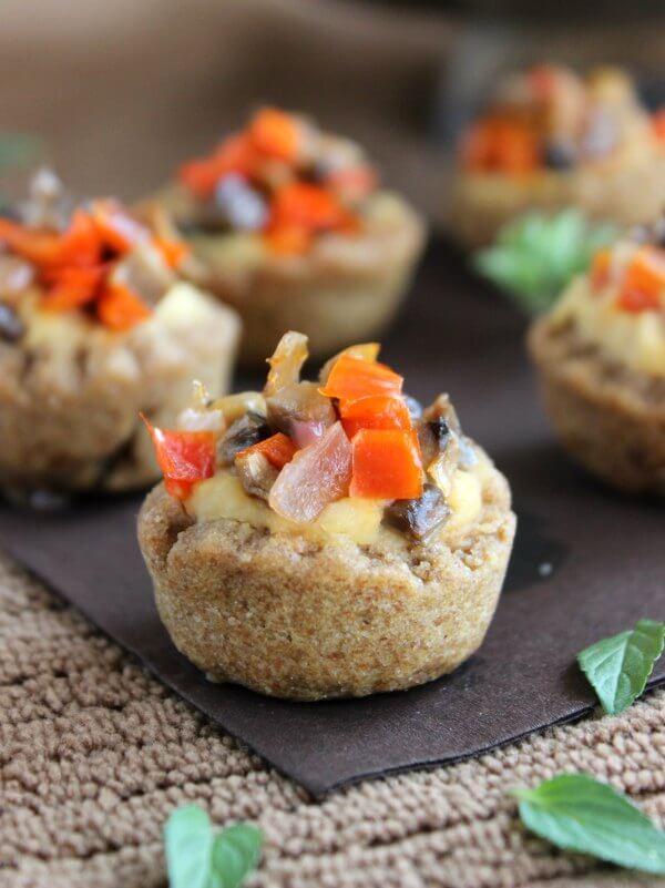 Garlic Hummus Stuffed Mini Bread Bowl is a scalloped baked whole wheat cup sitting on shades of brown. Filled with hummus and sprinkled with sauteed mushrooms and red bell peppers.