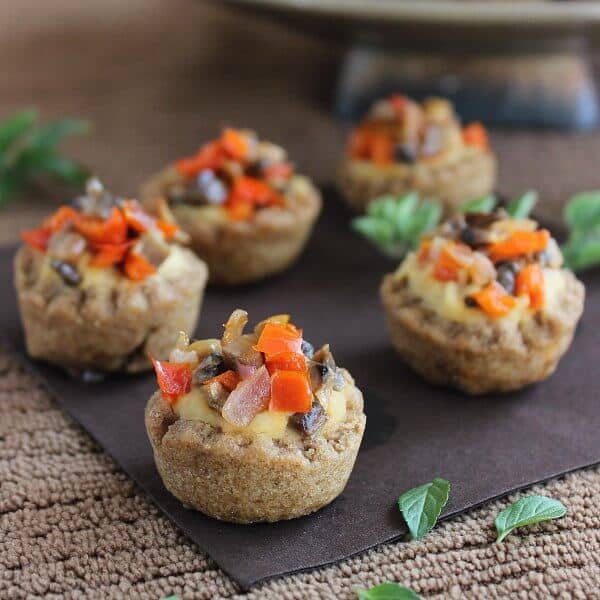 Garlic Hummus Stuffed Mini Bread Bowl is a scalloped baked whole wheat cup. Five cups are photographed on an angles and each is filled with hummus and sprinkled with sauteed mushrooms and red bell peppers..