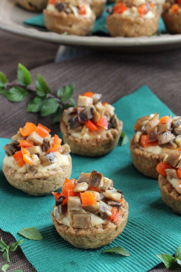 Garlic Hummus Stuffed Mini Bread Bowl is a scalloped baked whole wheat cup. Tilted cups are photographed to show them filled with hummus and sprinkled with sauteed mushrooms and red bell peppers.