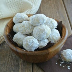 Treats covered with powdered sugar and are spilling over out of their brown wooden bowl and onto chocolate napkins.