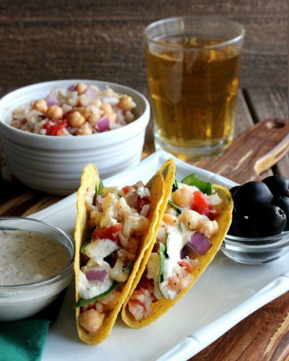 Ranch Cauliflower Tacos are angled toward the camera so there's a great view of corn tortillas filled with chunks of veggies and surrounded by homemade ranch dressing, black olives and a beer.