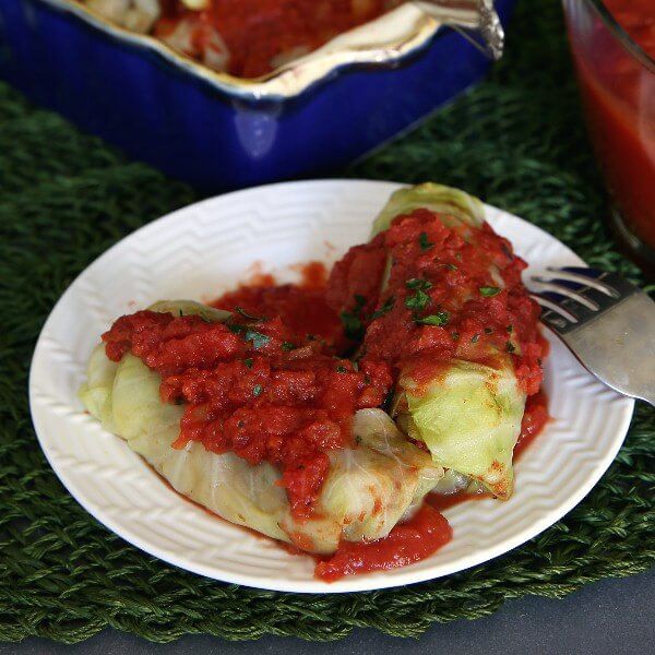 Vegan Cabbage Rolls Recipe is steamed cabbage rolled around a flavorful mushrooms, rice and lentils. Two are sitting on a white plate and topped with a rich red sauce.