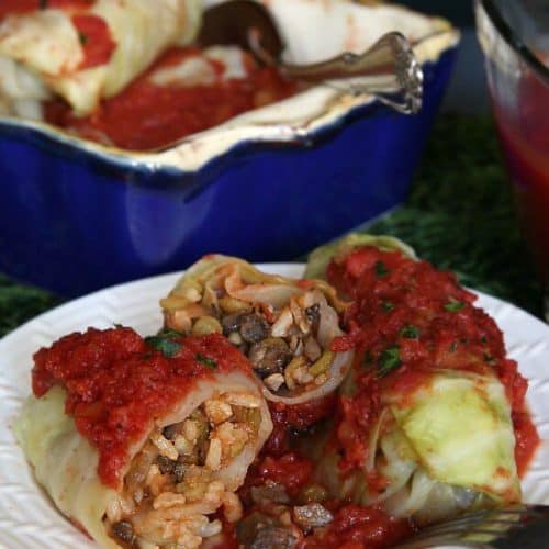 Vegan Cabbage Rolls Recipe is cabbage rolled around a flavorful mushrooms, rice and lentils. Two rolls are set on a white plate and cut open to show the tomato topped mix.