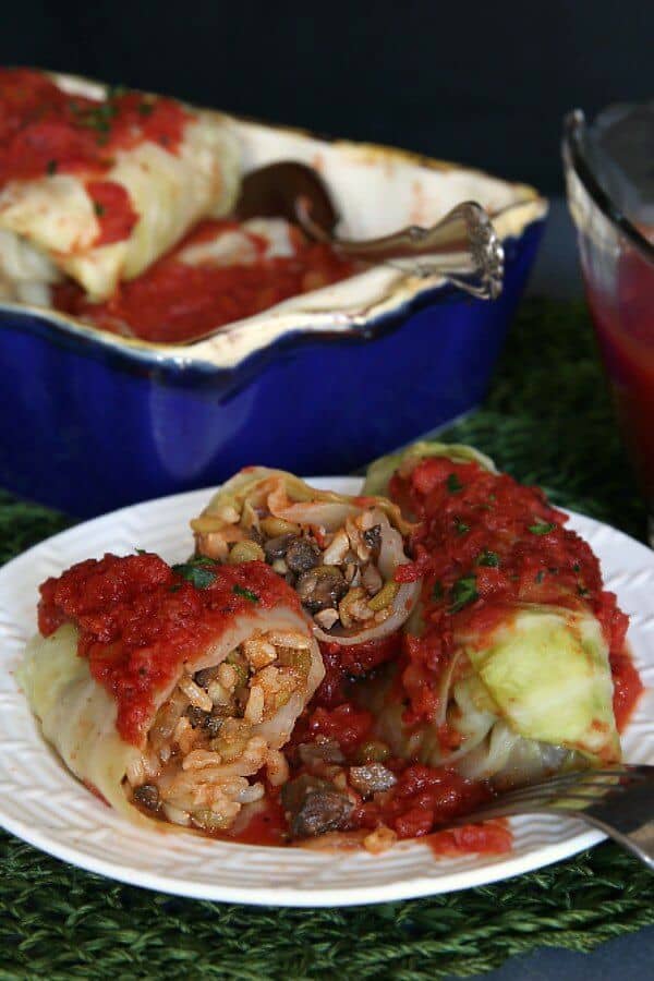 Vegan Cabbage Rolls Recipe is cabbage rolled around a flavorful mushrooms, rice and lentils. Two rolls are set on a white plate and cut open to show the tomato topped mix.