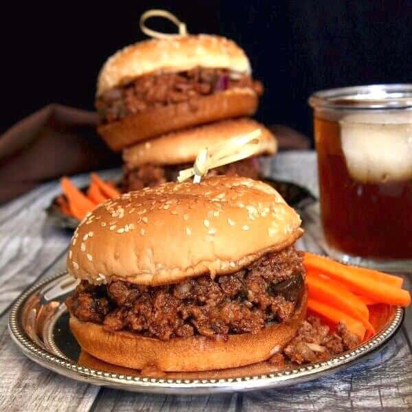 Vegan Sloppy Joes are stacked two high behind and one in front and all are perched on a silver tray. Bright carrot sticks are near at hand too.