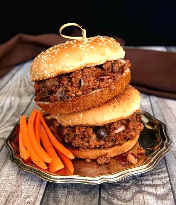 Vegan Sloppy Joes are stacked two high and perched on a silver tray. Bright carrot sticks are near at hand too.
