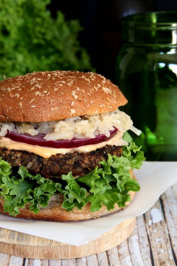 Crispy Anasazi Bean Burger is sitting on a square piece of parchment paper and the photo has cut the burger in half. green curly leaf lettuce, crispy brown burger and creamy colored sauerkraut finish the look.