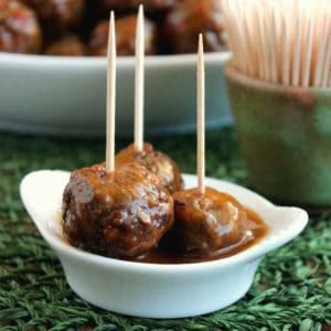 Vegan Lentil Balls Sweet and Sour Sauce Recipe has a closeup of three lentil balls sitting in a tiny white bowl of sweet and sour sauce. Tilted forward and each has a toothpick waiting to be taken.