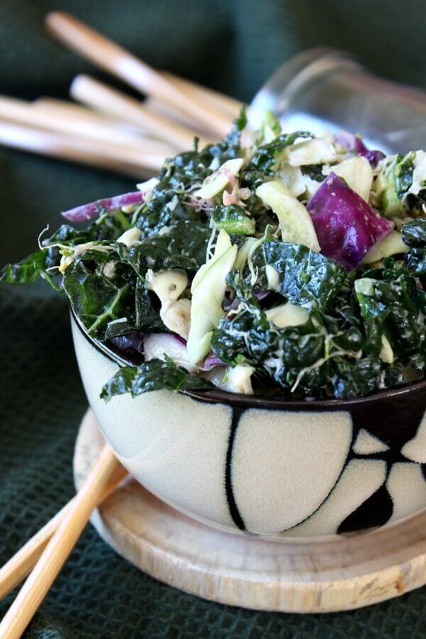 Massaged Kale Salad with Lemon Tahini Dressing is piled high in a bamboo designed bowl and shows green, white and purple veggies glistening with lemon tahini dressing.