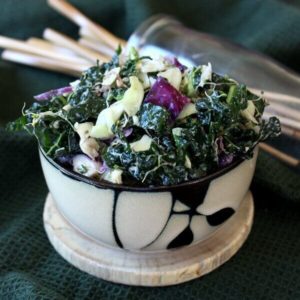 Massaged Kale Salad with Lemon Tahini Dressing is piled high in a bamboo designed bowl and tilted forward. Glistening tender greens, alfalfa shoots and contrasting purple and green cabbage is poking through.