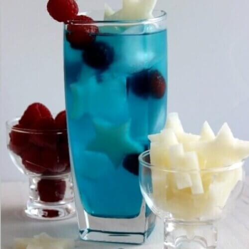 Patriotic Passion Cocktail is tall and blue with small white jicama stars and red raspberries floating inside.