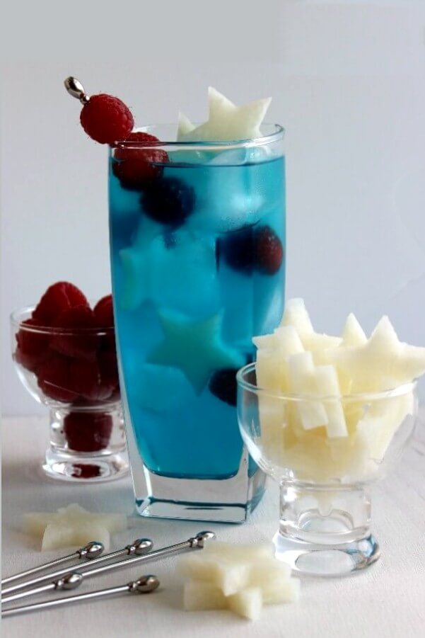 Patriotic Passion Cocktail is tall and blue with small white jicama stars and red raspberries floating inside.