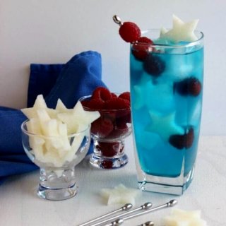 Patriotic Passion Cocktail is tall and blue with small white jicama stars and red raspberries sitting alongside a navy cloth napkin.