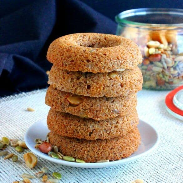 Baked Trail Mix Cake Donuts are stacked five high. Light golden brown and sitting on a white plate with hints of pumpkin seeds and nuts peeking out. A jar of trail mix sits behind.