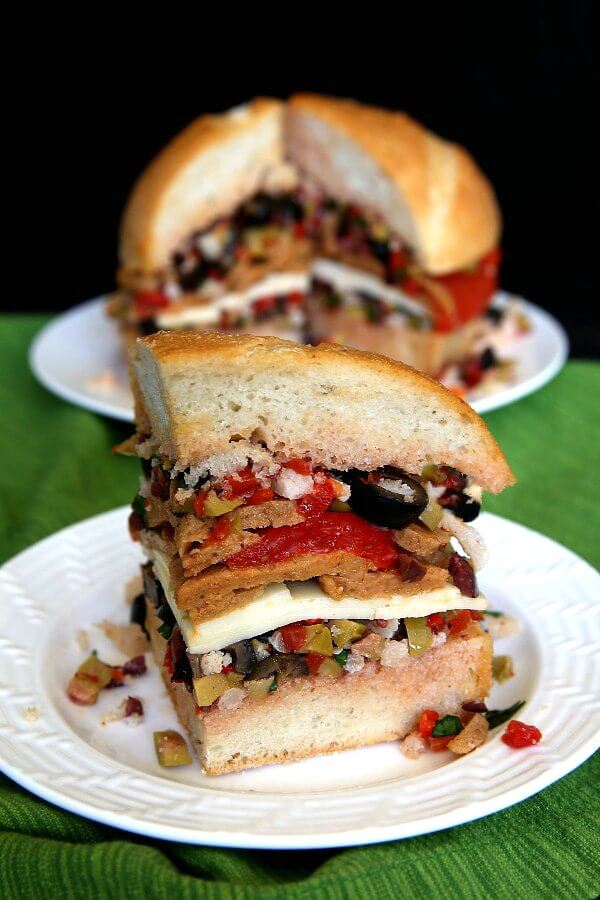 Vegan Muffuletta Sandwich is layers of mixed olives, roasted bell pepper, sliced seitan and dairy free cheese. Close-up front triangle slice that shows all the layers.side wedge slice just waiting for a big bite.