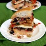 Vegan Muffuletta Sandwich is layers of mixed olives, roasted bell pepper, sliced seitan and dairy free cheese. Side wedge slice spilling out onto the white plate and just waiting for a big bite.