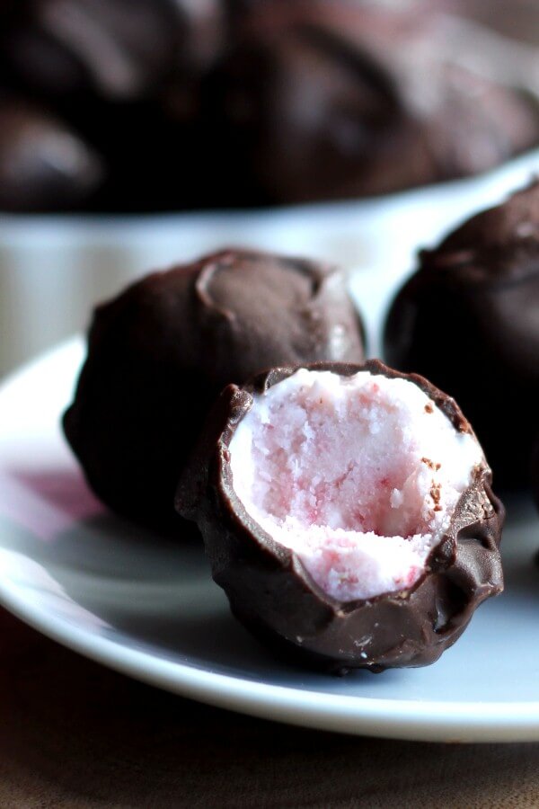 Dairy Free Strawberry Ice Cream Bonbons are sitting on a white plate and are three photographed to fill the whole frame. Covered in chocolate and one is broken open to show strawberry tinted ice cream.