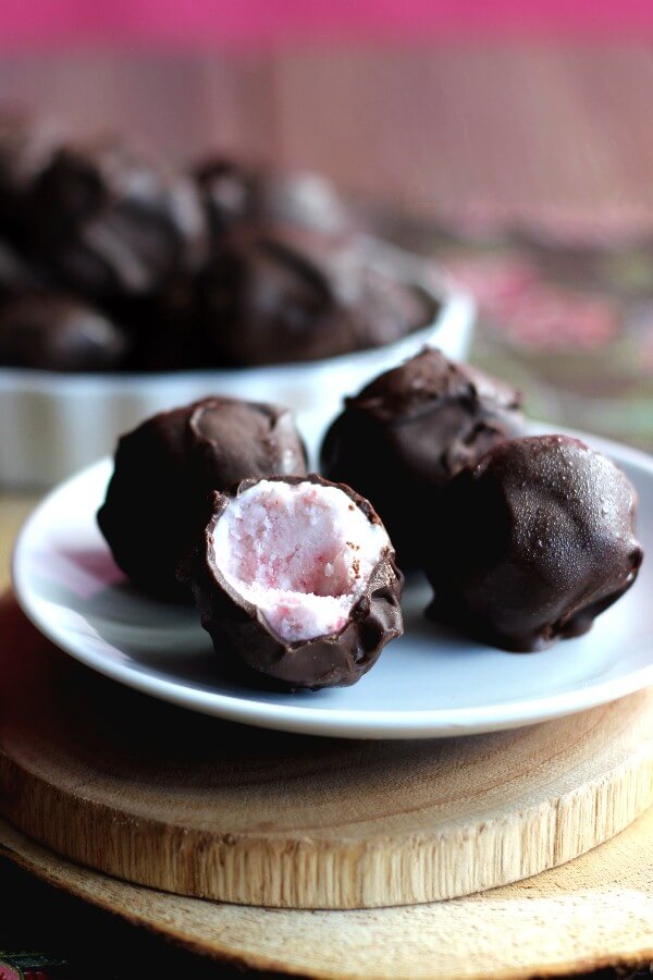 Dairy Free Strawberry Ice Cream Bonbons are sitting on a white plate. Three are covered in chocolate and one is broken open to show strawberry tinted ice cream.