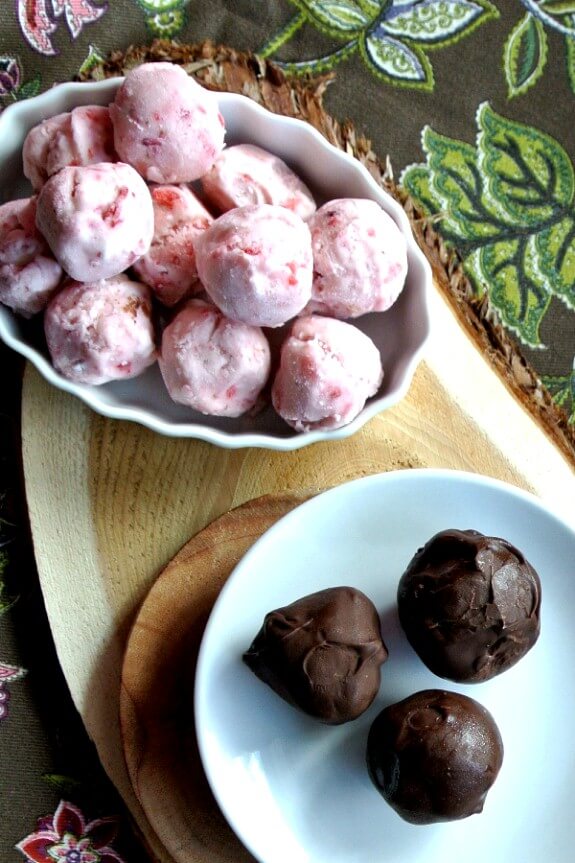 Dairy Free Strawberry Ice Cream Bonbons are sitting by three on a tiny white plate. A bowl of un-dipped strawberry ice cream balls are sitting and waiting to be dipped.