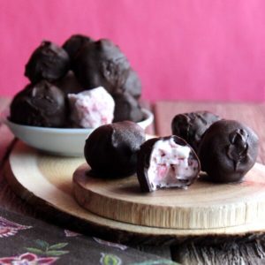 Dairy Free Strawberry Ice Cream Bonbons are sitting on a wooden disc. Three are covered in chocolate and one has been bit in half to show strawberry tinted ice cream.
