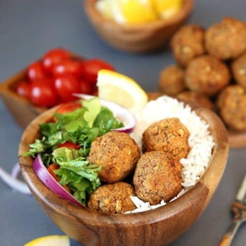 Baked Lentil Balls with Zesty Rice are piled in a wooden bowl next to a green salad and rice. Lemon slices and red onion slivers add color.