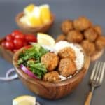Baked Lentil Balls with Zesty Rice are piled in a wooden bowl next to a green salad and rice. Lemon slices, red onion slivers and cherry tomatoes add color.
