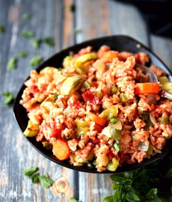 Garden Vegetable Rice Pilaf is is sharing a black bowl with green, red and orange vegetables. Sprinkled with fresh parsley with a spoon plunged in for an ample serving.