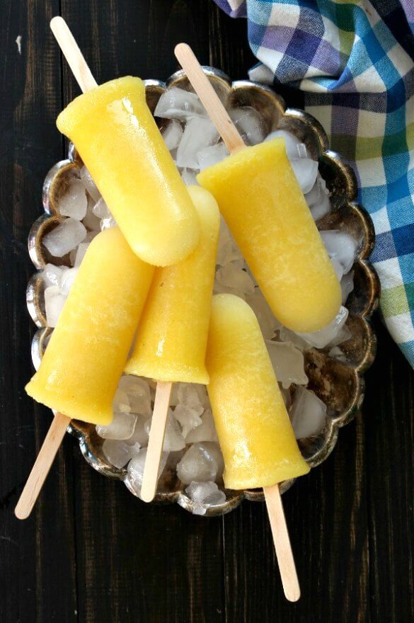 Pineapple Kiwi Popsicles are laying on a bed of ice in a silver tray. Five bright yellow popsicles and pointing every which way.