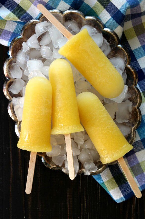 Pineapple Kiwi Popsicles are laying on a bed of ice in a silver tray. Four bright yellow popsicles with a blue and yellow check cloth under plate.