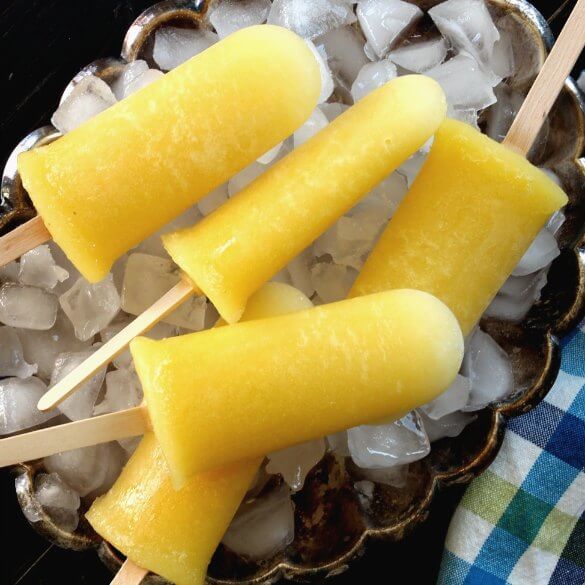 Pineapple Kiwi Popsicles are laying on a bed of ice in a silver tray. Five bright yellow popsicles and pointing every which way with a close-up.