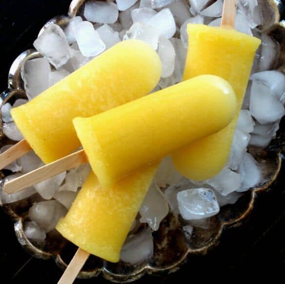 Pineapple Kiwi Popsicles are laying on a bed of ice in a silver tray. Four bright yellow popsicles and pointing every which way.