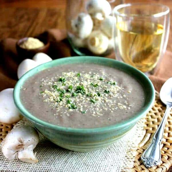Best Creamy Mushroom Soup is filling a green stripped pottery bowl with vegan parmesan and parsley sprinkled on top. Glass of white wine behind.