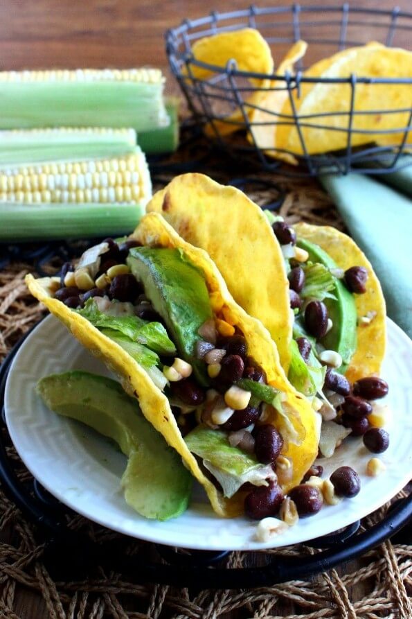 Corn and Black Bean Salsa Tacos are two to a plate and tilted forward. The shells are overflowing with green, yellow and black beans. White plate is sitting on a black trivet.