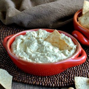 Baba Ghanoush Dip is loaded in a red handled bowl with p-ita chip poking inside and waiting to be scooped up. Sitting on a chocolate beaded mat.