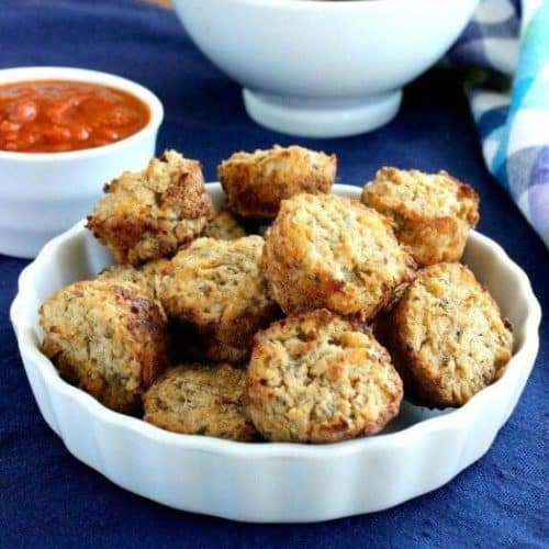 Cauliflower Pizza Bites are little mini muffins and are piled high in a white scalloped bowl on a cobalt blue placemat. Pizza sauce and a blue checked napkin on the side.