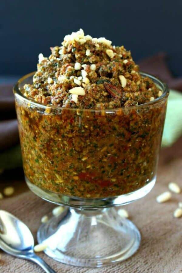 Vegan Sun Dried Tomato Pesto is in a clear glass footed container. Rich reds piled high with a spoon on the side.