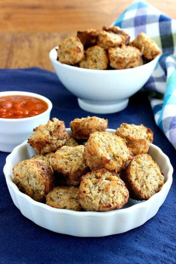 Cauliflower Pizza Bites are little mini muffins and are piled high in a white scalloped bowl on a cobalt blue placemat. Pizza sauce in a small bowl on the side and a big bowl of more muffins behind.