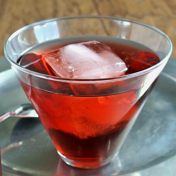 Pomegranate Swizzle is bright red and showing through a flared glass with a giant ice cube floating inside. Sitting on a pewter tray with a silver swizzle spoon sitting next to it.