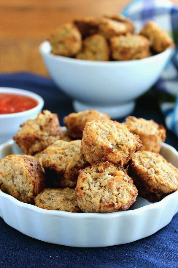 Cauliflower Pizza Bites are little mini muffins and are piled high in a white scalloped bowl on a cobalt blue placemat. Pizza sauce in a small bowl on the side.