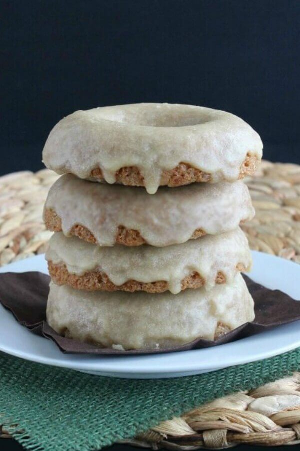 Breakfast doughnuts are stacked four high with each having a maple glaze dripping over the sides. Sitting on a white plate and a green burlap square.