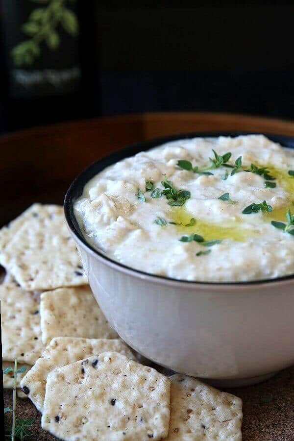 Chunky Chickpea Dip is in an ivory bowl with a black interior to show off the dip. Crackers are on the side and fresh herbs are sprinkled on the hummus.