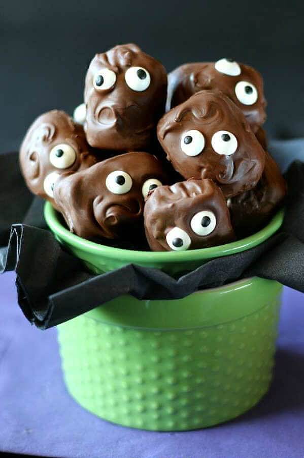 Copycat Almond Joy Candy Bars are piled in a green jar with their googly eyes looking at you. Swirly chocolate hair and mouths are in scary and cuts poses.