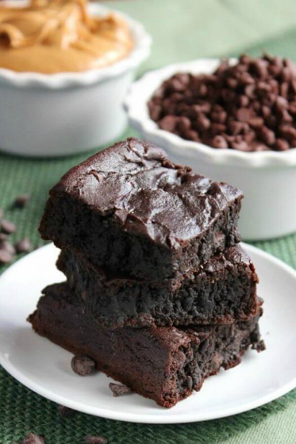 Crazy Brownies are cut into squares and stacked three high on a small white plate and green burlap mat. Chocolate chips are in a white bowl behind.