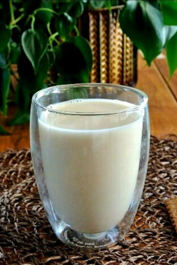 Homemade Almond Milk is filling a double walled clear glass tumbler. Sitting on a forest green mat with a houseplant behind.