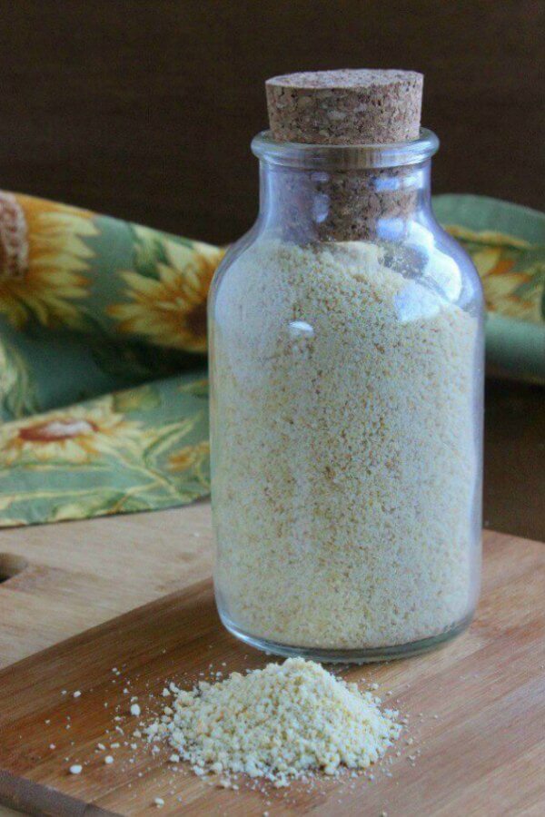 Vegan Parmesan Cheese is in a clear cork bottle with a pile spilled in front. All is on a wooden cutting board with sunflower cloth napkin behind.