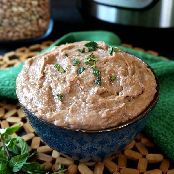 Instant Pot Refried Beans are piled high in a blue bowl and sprinkle with fresh herbs. It's sitting on a open wood mat and tilted towards you and a green cloth laying behind.