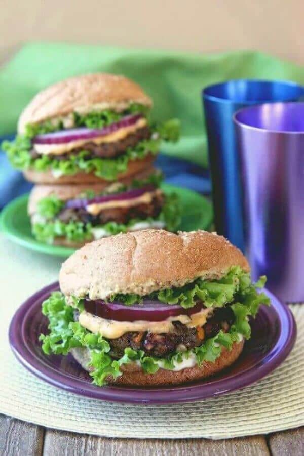 Kidney and Black Bean Burgers are on a big bun with layers rippled lettuce, patty creamy orange sauce, a big fat red onion slice and more. All on a purple plate.