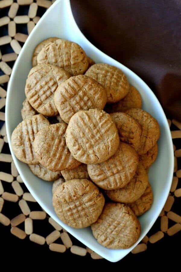 Maple Peanut Butter Cookies are seen with an overhead photo. The cookies are piled in a swirl shaped white bowl and places on an open-weave wooden mat.