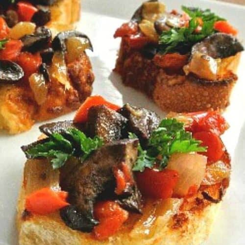 Mushroom Bruschetta Crostini is a mix of mushrooms, onions and red bells. The mix is piled high on small toasts and plated on a white square plate.