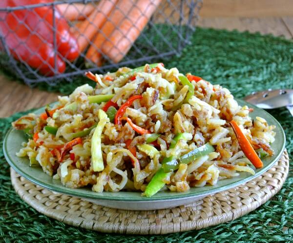 Rainbow Veggie Hashbrown Potatoes are fried together with carrots, bells, zucchini and onions. Pilerd high in a green and white bowl on a green mat. Fresh Red bells and carrots are behind in a wire basket.